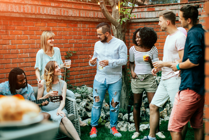 Happy young people at the party in the backyard talking and smiling. One young man playing guitar. They are enjoy spending time together.