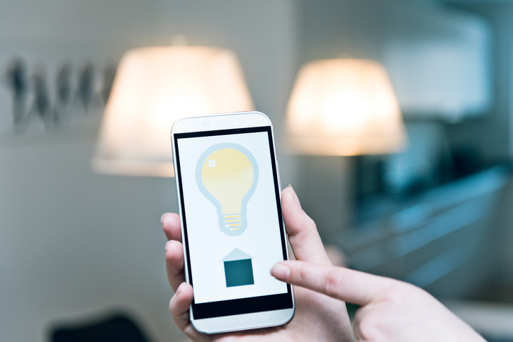 Smartphone is used to control the lights in a home. Woman holds a phone up in front of two lights hanging from the ceiling, The screen shows an icon for a light blub and the possbility for turning the lights at home on and off. It is a concept of internet of things with a mobile phone. Note: The image on the screen is my work, not a functional app.